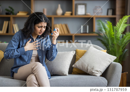 Sick young Indian woman sitting on sofa at home, holding her hand on her chest, suffering from asthma, using inhaler. Sick young Indian woman sitting on sofa at home, holding her hand on her chest, suffering from asthma, using inhaler. 131787959