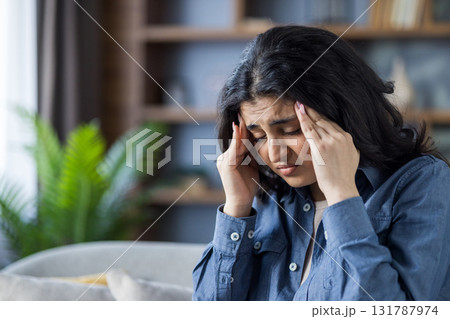 Close-up photo of a young Indian woman sitting cross-legged on a sofa, holding her head in her hands, getting a massage, feeling severe pain and pressure. 131787974