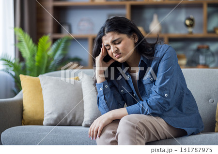 Suffering from headache and pressure, Indian young woman sits on sofa at home and grimaces holding her head. Suffering from headache and pressure, Indian young woman sits on sofa at home and grimaces holding her head. 131787975