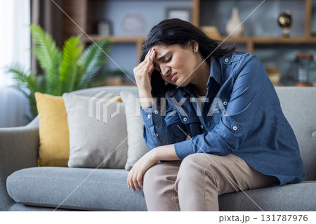 A young woman is experiencing a headache while sitting on a sofa at home, looking stressed. She is clutching her forehead. A young woman is experiencing a headache while sitting on a sofa at home, looking stressed. She is clutching her forehead. 131787976