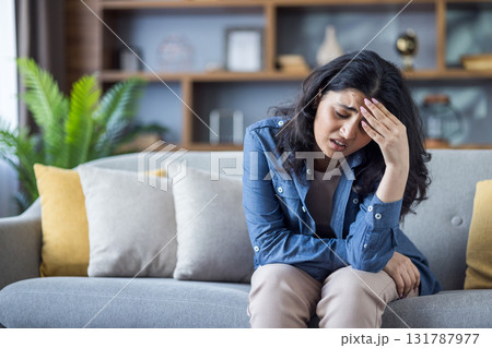 Tired young Indian woman in denim shirt sitting on sofa at home and holding her head, feeling severe pain and tension. Tired young Indian woman in denim shirt sitting on sofa at home and holding her head, feeling severe pain and tension. 131787977