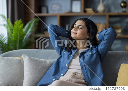 A serene young woman relaxes on a comfortable couch, hands behind her head, and closed eyes. She embodies tranquility in a cozy, home setting. 131787998