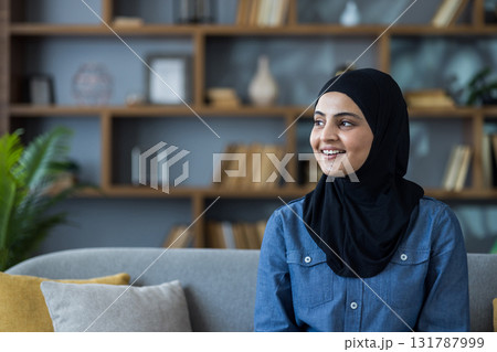 Close-up portrait of a young Muslim woman in a hijab and shirt sitting on a sofa at home, smiling and looking to the side. 131787999