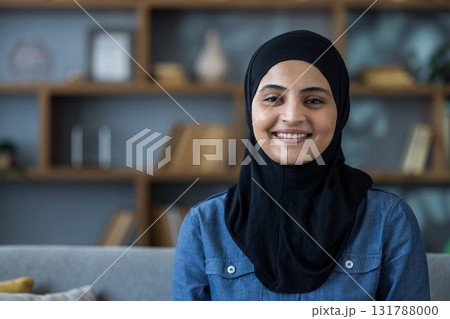 Close-up portrait of a young Muslim woman in a black hijab sitting on a sofa at home and smiling at the camera. Close-up portrait of a young Muslim woman in a black hijab sitting on a sofa at home and smiling at the camera. 131788000