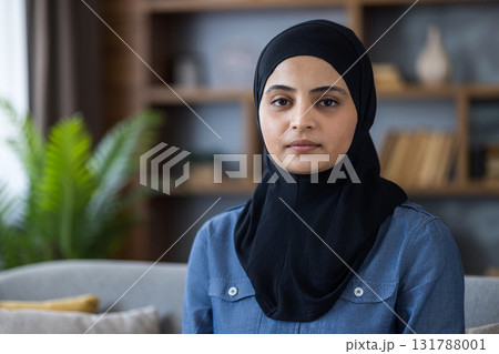 A portrait of a serene muslim woman wearing a black hijab. She is sitting inside and looking straight at the viewer. 131788001