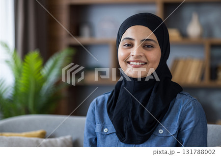 Portrait of a young smiling Muslim woman in a black hijab at home and looking at the camera. Portrait of a young smiling Muslim woman in a black hijab at home and looking at the camera. 131788002