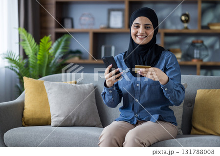 Smiling young Muslim woman in hijab sitting on sofa at home, holding credit card and phone, looking at camera. Smiling young Muslim woman in hijab sitting on sofa at home, holding credit card and phone, looking at camera. 131788008