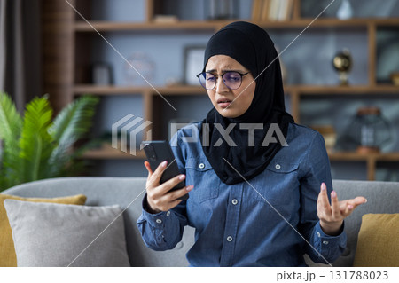 Worried young Muslim woman in hijab sitting on sofa at home, looking at mobile phone screen and spreading her hands in frustration. 131788023