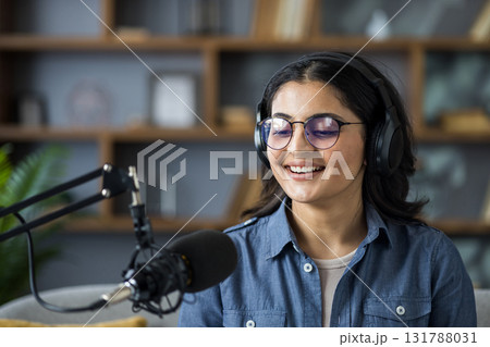 Close-up photo of a young Indian woman wearing glasses and headphones sitting at home and talking into a microphone stand. Close-up photo of a young Indian woman wearing glasses and headphones sitting at home and talking into a microphone stand. 131788031
