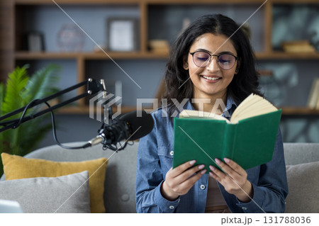 Close-up photo of a young Indian girl at home, sitting on the couch and reading into a microphone from a book she is holding. 131788036