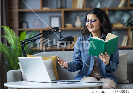 Smiling young Indian woman sitting on sofa at home, holding a book, reading and talking on laptop camera via video call. 131788037