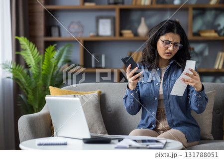 Upset and disappointed young Indian woman sitting on the couch at home, holding her phone and checking bills and receipts. 131788051