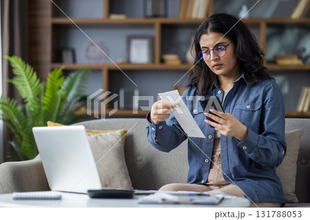 Upset young Indian woman sitting on sofa at home near table with laptop, holding phone in hands and looking worriedly at receipts and bills. Upset young Indian woman sitting on sofa at home near table with laptop, holding phone in hands and looking worriedly at receipts and bills. 131788053
