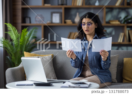 Worried young Indian woman sitting on the couch at home and looking shocked at the documents she is holding. Worried young Indian woman sitting on the couch at home and looking shocked at the documents she is holding. 131788073