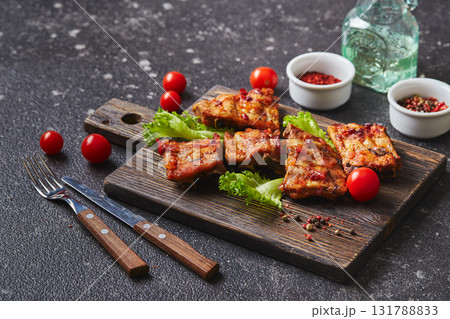 Grilled baked pork ribs with spices and vegetables on wooden cutting board on dark background. American food concept. Grilled baked pork ribs with spices and vegetables on wooden cutting board on dark background. American food concept. 131788833