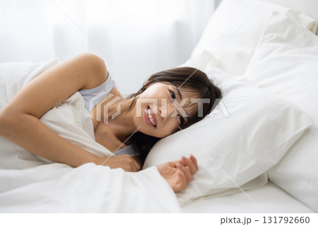 朝の光に包まれて白いベッドで微笑む若い女性 朝の光に包まれて白いベッドで微笑む若い女性 131792660