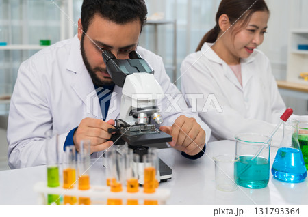 Male scientist with beard using microscope in laboratory wearing white lab coat while female colleague working among various colorful beakers and test tubes 131793364