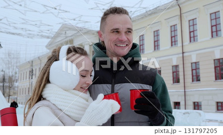 Cheerful couple sipping warm beverages amid snowflakes, winter urban square decorated festively, radiating seasonal happiness and intimate connection Cheerful couple sipping warm beverages amid snowflakes, winter urban square decorated festively, radiating seasonal happiness and intimate connection 131797205