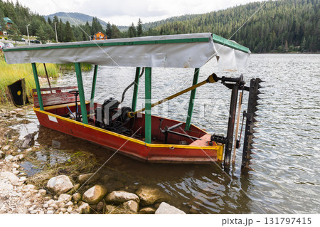 A bright red boat with a green frame and white canopy rests at a rocky lakeside 131797415