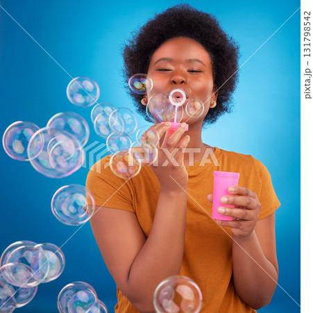 Fun, bubbles and black woman with joy, positive attitude and happiness against a blue studio background. African American female, lady and soap bubble for joy, playing and cheerful with excitement 131798542