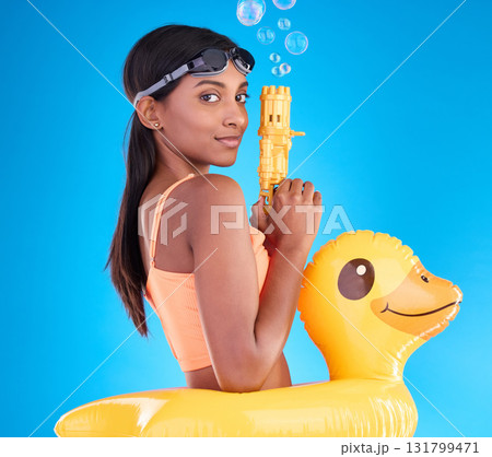 Portrait, bubbles and rubber duck with a woman on a blue background in studio ready for summer swimming. Happy, travel and vacation with an attractive or playful young female holding a bubble gun 131799471
