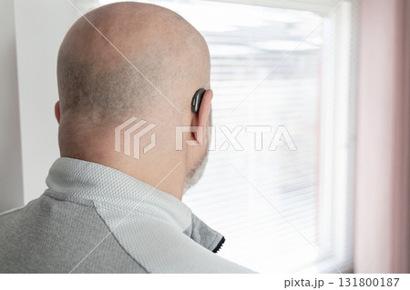 A bald man with a hearing aid looks out the window A bald man with a hearing aid looks out the window 131800187