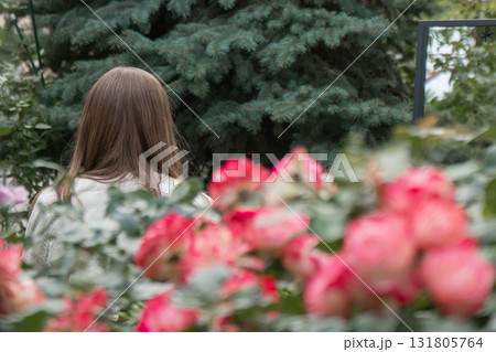 A woman with long brown hair stands in a blooming garden, her back turned toward the viewer, framed by pink roses and evergreen trees 131805764
