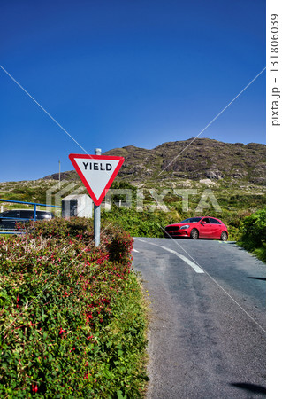 A red car drives on a scenic road in Ireland with a yield sign. A red car drives on a scenic road in Ireland with a yield sign. 131806039