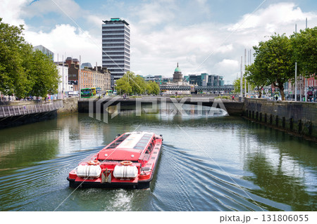 A vibrant shot of Dublin, Ireland, capturing the River Liffey with a tourist boat, bridges, and cityscapes under a bright sky. 131806055