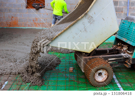 Wet concrete is being poured from dumping wheeled concrete wheelbarrow into foundation area at construction site 131806418