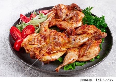 Baked quail, served on a plate with crispy lettuce leaves, fresh tomatoes and herbs, on a dark plate, homemade, 131808235