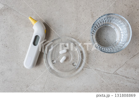 White digital thermometer for temperature, pill and glass with water on the stone table. Healthcare from pharmacy. Person sit at home because of illness. White digital thermometer for temperature, pill and glass with water on the stone table. Healthcare from pharmacy. Person sit at home because of illness. 131808869