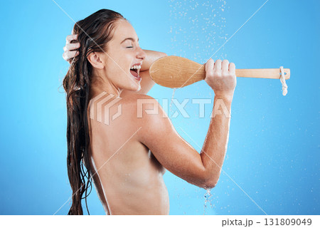 Shower singing, skincare and woman with happiness karaoke in a studio. Bathroom, music and happy wet female model with isolated blue background and cleaning brush for beauty routine, wellness and fun 131809049