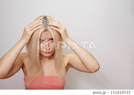 Emotive portrait of a blonde woman holding her head with both hands, expressive face and dramatic pose. Regrowth of hair roots, gray hair, hair problems, hair loss or dandruff of scalp. Beauty concept 131810535