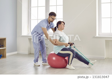 Nurse man helping fat patient woman to do stretching exercises with rubber band sitting on fit ball 131813047