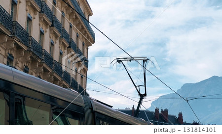 Close-up view of a modern tram passing through Grenoble, France. Historic building facades, power lines, and alpine mountains visible in the background 131814101