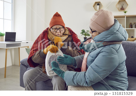 Senior family couple wearing winter clothes warming up by electric heater at home Senior family couple wearing winter clothes warming up by electric heater at home 131815329