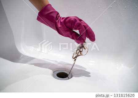 Hand of woman in pink rubber glove taking off hair clump from water drain, blocked pipe in bath. 131815620