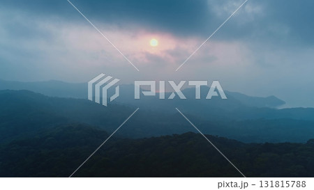 Dark green forested hills and mountains fading into the misty distance with sun setting behind clouds illuminating sky with soft orange and pink light 131815788