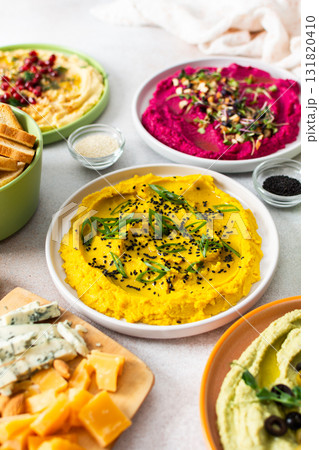 A variety of hummus flavors including turmeric, classic, and broccoli are served with fresh bread, nuts, and garnishes on a colorful charcuterie board, perfect for sharing A variety of hummus flavors including turmeric, classic, and broccoli are served with fresh bread, nuts, and garnishes on a colorful charcuterie board, perfect for sharing 131820410