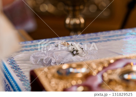 Intimate Wedding Ring Ceremony Detail: Gold Bands on White Pillow with Lace, Floral Accent in Church Setting. 131829525