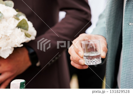 Groom Holding Shot Glass at Wedding, Bouquet and Suit Details, Soft Focus, Brown and Gray Formal Attire, Celebration Tradition. 131829528