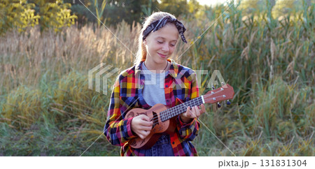 cute girl playing acoustic ukulele guitar outside grass happy portrait. a happy, young girl playing an acoustic guitar or ukulele outside on the grass to convey music, fun, and a cute lifestyle 131831304