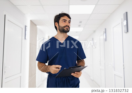 A doctor in blue scrubs walks down a brightly lit hospital hallway, holding a clipboard and pen, likely reviewing patient information. A doctor in blue scrubs walks down a brightly lit hospital hallway, holding a clipboard and pen, likely reviewing patient information. 131832732