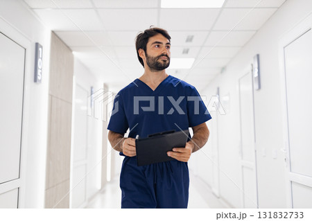 A doctor in scrubs walks down a bright hallway, holding a clipboard, likely heading to see a patient. A doctor in scrubs walks down a bright hallway, holding a clipboard, likely heading to see a patient. 131832733