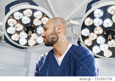 A surgeon stands in an operating room, illuminated by bright surgical lights, ready to perform a procedure. 131832756