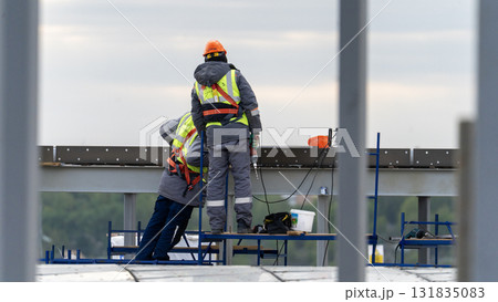 Construction workers in their work attire are installing fresh roofing components, power tools, and electric drills, employing them on the new rooftops of towering buildings. 131835083