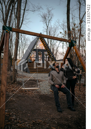 Couple enjoys a quiet moment on a swing in front of a cozy cabin in the woods during dusk 131837422