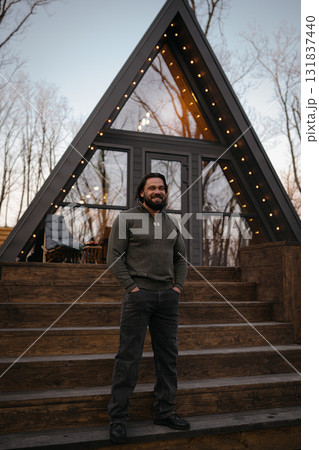Man stands on steps of modern cabin in forest during evening light Man stands on steps of modern cabin in forest during evening light 131837440