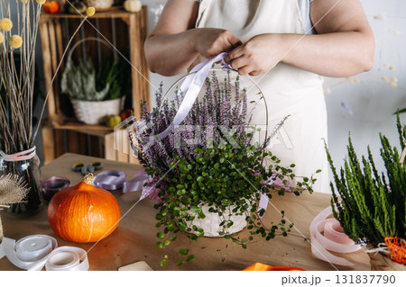 Person tying lavender ribbon around floral gift basket filled with heather and trailing greenery on a rustic table. Thanksgiving bouquet, handmade gift basket, seasonal flower gift, fall ribbon decor Person tying lavender ribbon around floral gift basket filled with heather and trailing greenery on a rustic table. Thanksgiving bouquet, handmade gift basket, seasonal flower gift, fall ribbon decor 131837790